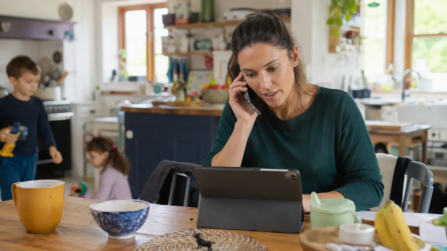 A woman speaking by phone and checking a tablet at home while children play